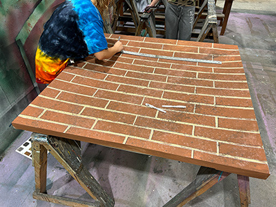 A student wearing a tie-dye shirt works on a painted surface designed to look like a brick wall. The large, textured board rests on sawhorses, and chalk lines and a ruler are visible on the surface, showing the process of creating realistic brick patterns.