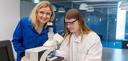 A woman with blond hair instructing a student on a scientific experiment in a lab.